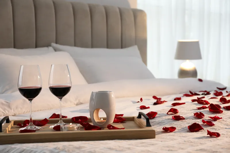 Tray with glasses of red wine, candles and rose petals on bed in room
