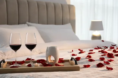 Tray with glasses of red wine, candles and rose petals on bed in room