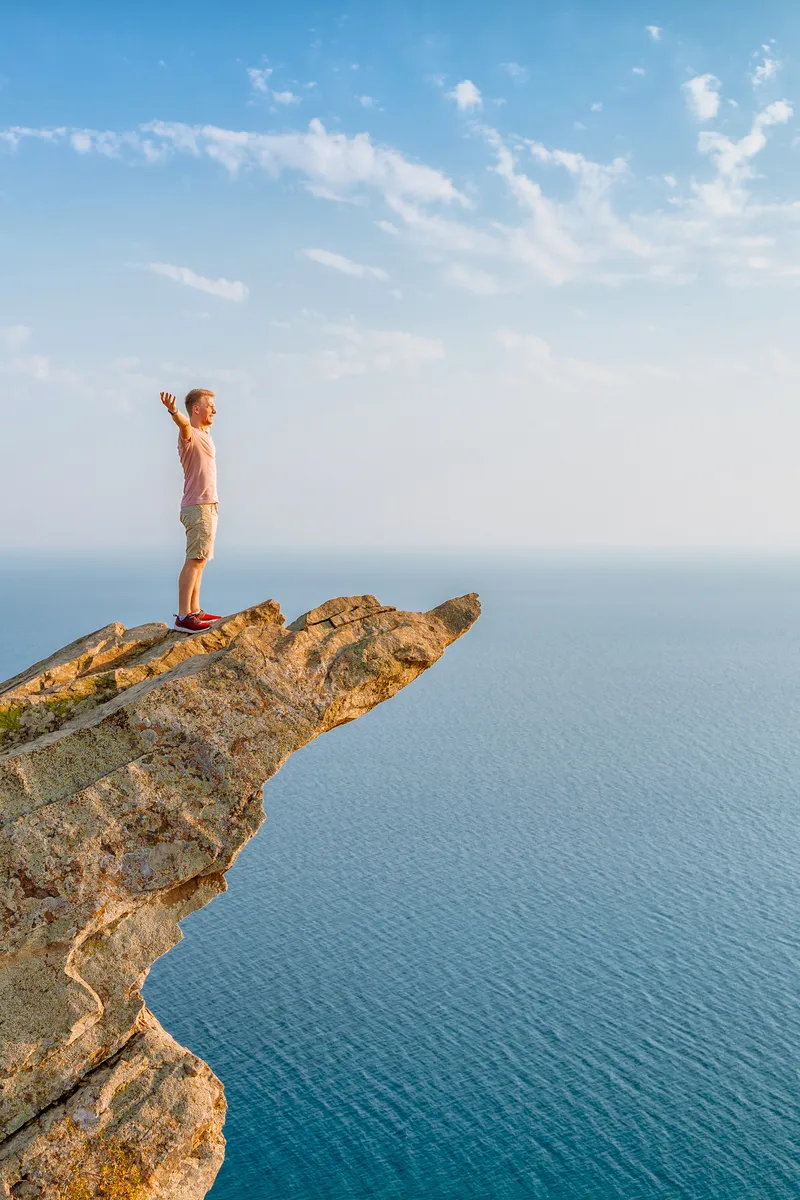 A young athletic man stands on a picturesque steep cliff above the sea against the sky in Crimea. The concept of travel and freedom.
