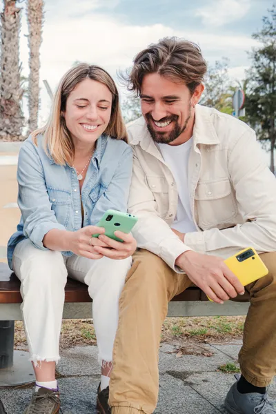 Vertical portrait of a young adult friends using their cellphones, browsing on internet enjoying free time sitting on bench outside. Happy caucasian couple having fun together with their smart phones