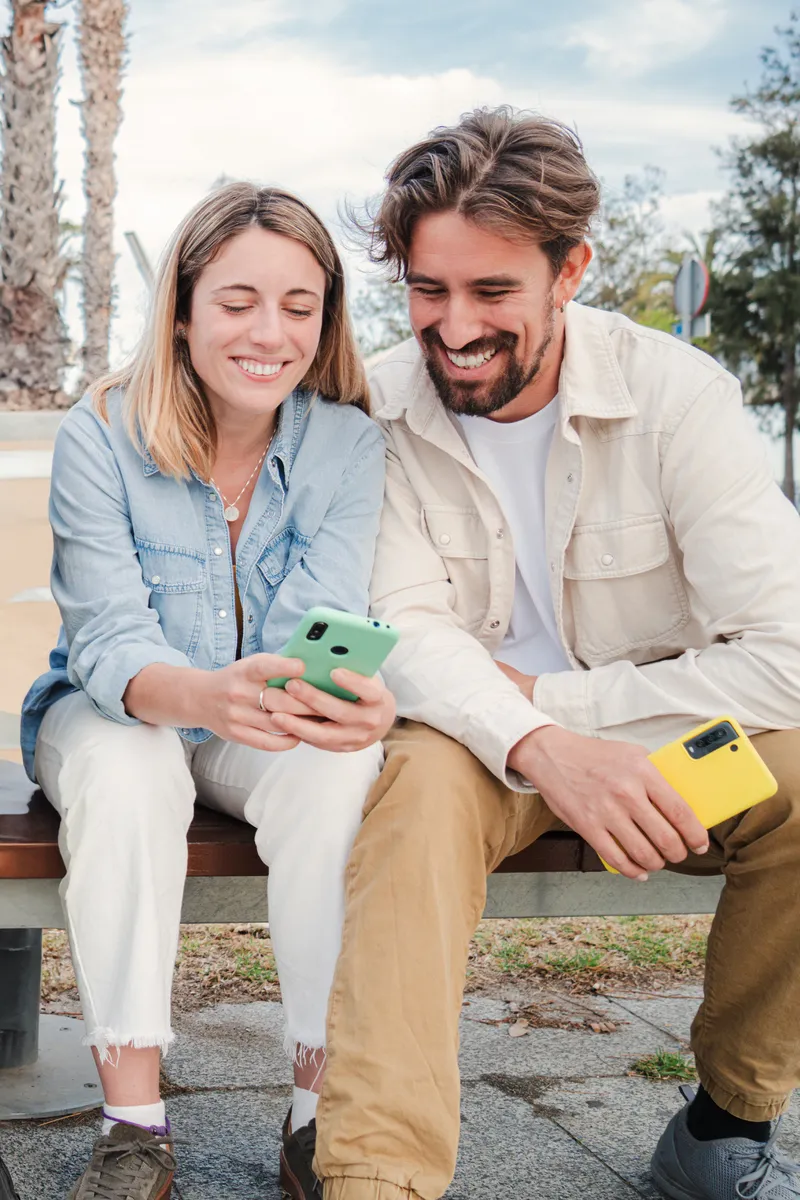 Vertical portrait of a young adult friends using their cellphones, browsing on internet enjoying free time sitting on bench outside. Happy caucasian couple having fun together with their smart phones