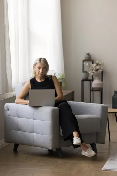 Serious mature woman checks email sitting on armchair with laptop